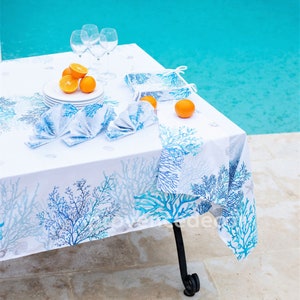 May include: White tablecloth with a blue coral reef pattern. The tablecloth is set with white plates, blue napkins, and oranges. There are also wine glasses on the table.