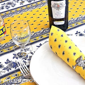 May include: A yellow and white floral patterned tablecloth featuring a Ricard bottle, a Ricard glass, a dark bottle of Pastis de Marseille, a silver fork, a white plate, and a yellow napkin held by a silver metal owl napkin ring.