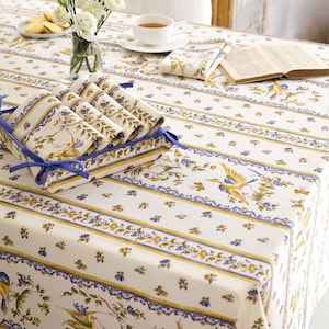May include: A white tablecloth with a blue and yellow floral pattern featuring birds. The tablecloth is set with a cup and saucer, a book, and a set of folded napkins.