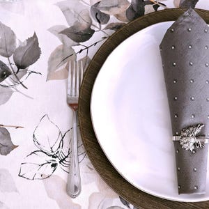 May include: A formal table setting featuring a white plate atop a wooden charger, with a gray napkin adorned with rhinestones and a decorative napkin ring. A silver fork rests on a floral patterned tablecloth.