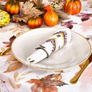 May include: A fall-themed table setting with a white plate, gold cutlery, and a folded napkin in a decorative ring. The tablecloth features a leaf pattern, and small pumpkins and gourds are arranged around the plate.
