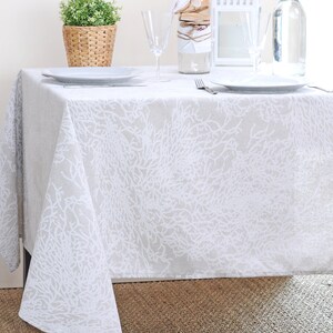 May include: A white tablecloth with a beige background and a white coral design. The tablecloth is laid out on a table with a white plate, a glass, and a fork on top.