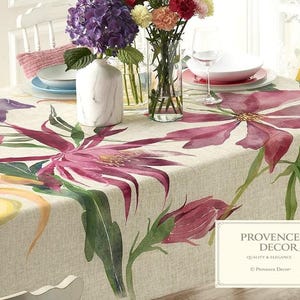 May include: A beige tablecloth with a large floral design in shades of pink, purple, and yellow. The table is set with white plates, wine glasses, and a vase of flowers. The tablecloth has the text "PROVENCE DECOR" in the corner.