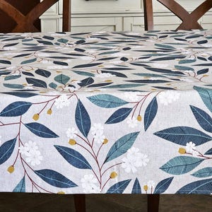 May include: A rectangular tablecloth with a botanical pattern. The design features dark blue and teal leaves, white flowers, and yellow accents on a light gray background. The tablecloth is draped over a wooden table.