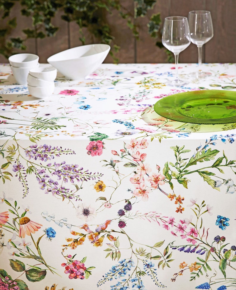 May include: White tablecloth with a floral pattern featuring pink, purple, yellow, and blue flowers. The tablecloth is set with white bowls and two wine glasses.