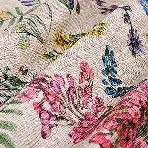 May include: A close-up of a beige fabric with a floral print. The fabric features a variety of flowers in shades of pink, blue, yellow, and green. The fabric has a textured surface, giving it a rustic look.