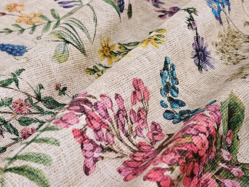 May include: A close-up of a beige fabric with a floral print. The fabric features a variety of flowers in shades of pink, blue, yellow, and green. The fabric has a textured surface, giving it a rustic look.