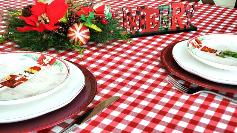 Pu&ograve; includere: Una tovaglia a quadri rossi e bianchi con un cartello di legno che dice "Merry". Ci sono due posti a tavola con piatti bianchi e sottopiatti rossi. Un centrotavola natalizio con una stella di Natale, verde e bastoncini di zucchero &egrave; sullo sfondo.
