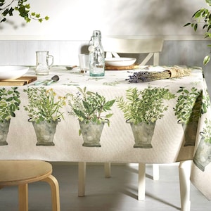 May include: A beige tablecloth with a green herb print. The tablecloth features a repeating pattern of potted herbs, including basil, rosemary, and thyme. The tablecloth is laid on a white table with a white chair and a wooden stool.