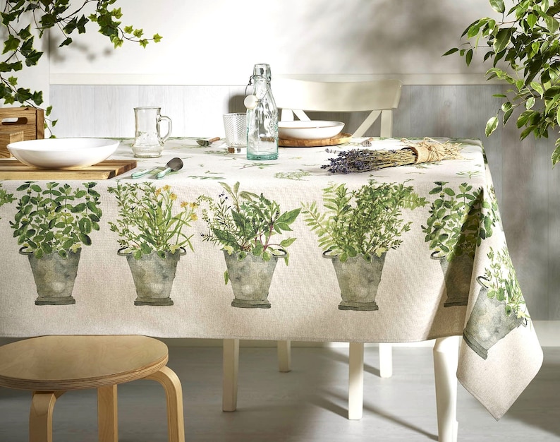 May include: A beige tablecloth with a green herb print. The tablecloth features a repeating pattern of potted herbs, including basil, rosemary, and thyme. The tablecloth is laid on a white table with a white chair and a wooden stool.
