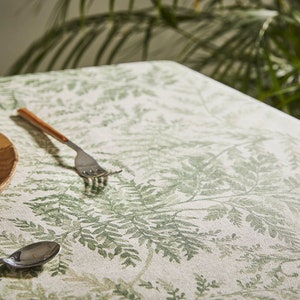 May include: A white tablecloth with a green fern pattern. A wooden plate with a bowl and a napkin is on the table. A fork and spoon are also on the table.