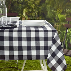 May include: A black and white checkered tablecloth covers a white table set for dining outdoors. White bowls and plates are on the table, along with glasses and a water pitcher. The background features green foliage and a wooden chair.