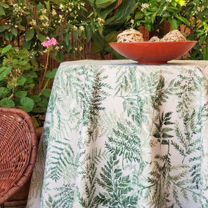 May include: A white tablecloth with a green fern pattern. The tablecloth is laid on a table with a brown wicker chair in the foreground.
