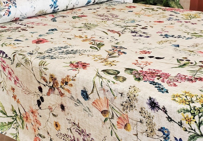 May include: A beige fabric with a floral pattern featuring various shades of pink, blue, yellow, and green flowers. The fabric has a subtle linen texture.
