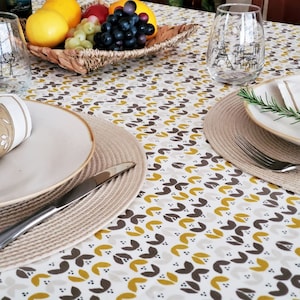 May include: A white tablecloth with a repeating pattern of brown and yellow leaves. Two place settings are visible, each with a white plate, a woven placemat, a silver knife, and a fork. A glass of water is on the table, and a bowl of fruit is in the background.