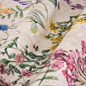 May include: A close-up of a beige fabric with a floral print. The fabric features a variety of flowers in shades of pink, blue, yellow, and green. The fabric has a textured surface, giving it a rustic look.