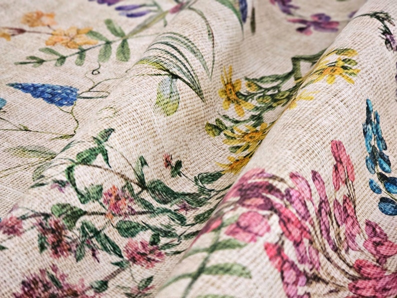 May include: A close-up of a beige fabric with a floral print. The fabric features a variety of flowers in shades of pink, blue, yellow, and green. The fabric has a textured surface, giving it a rustic look.