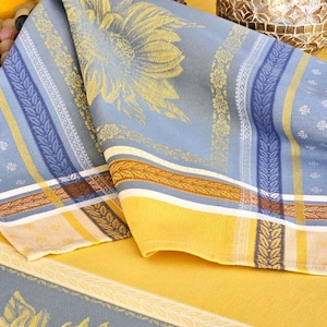 May include: A folded fabric napkin featuring a woven pattern of yellow sunflowers on a light blue background. The design includes vertical stripes in dark blue, white, and yellow, with intricate leaf motifs and small floral accents throughout the textile.