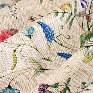 May include: A close-up of a beige fabric with a floral pattern. The fabric features a variety of flowers in shades of blue, pink, and green. The fabric has a textured surface, giving it a rustic look.