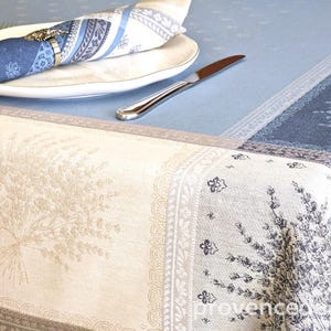 May include: A blue and cream tablecloth with a floral pattern, set with a white plate, a blue napkin, and a silver knife. The napkin is held by a silver ring. The tablecloth features a lavender sprig design.