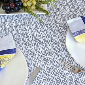 May include: Two place settings with white plates and blue and yellow patterned napkins. The napkins are folded into a triangle shape. The tablecloth is white with a blue and white geometric pattern.