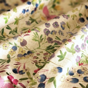 May include: Close-up of a cream-colored fabric with a floral pattern. The design features small, colorful flowers in shades of pink, purple, blue, and yellow, along with green leaves. The fabric is slightly wrinkled, suggesting a soft texture.