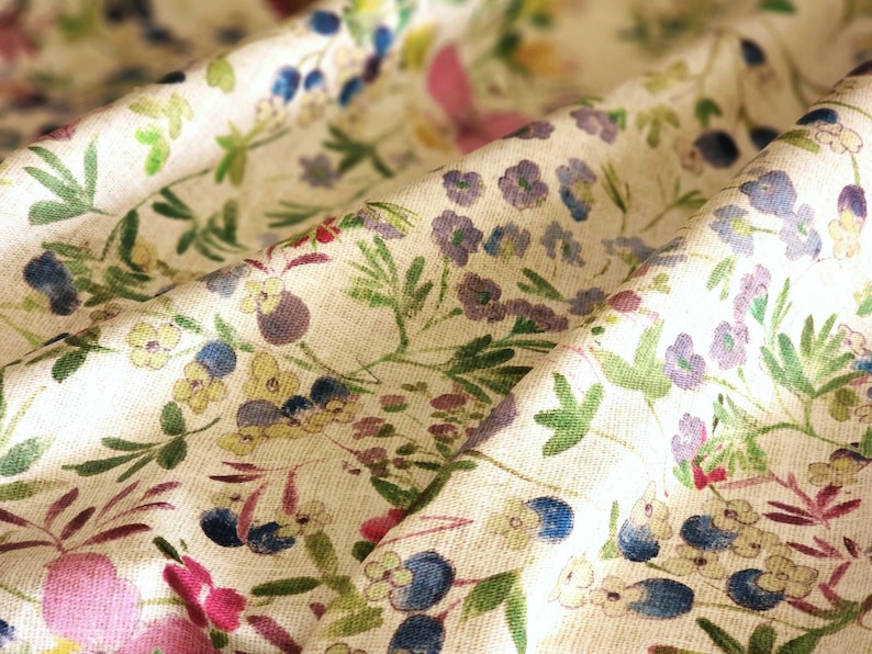 May include: Close-up of a cream-colored fabric with a floral pattern. The design features small, colorful flowers in shades of pink, purple, blue, and yellow, along with green leaves. The fabric is slightly wrinkled, suggesting a soft texture.