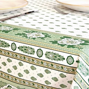 May include: A white and green floral tablecloth with a detailed border featuring paisley and floral patterns. The tablecloth has a repeating pattern of small green leaves. A knife and fork are visible on the table.