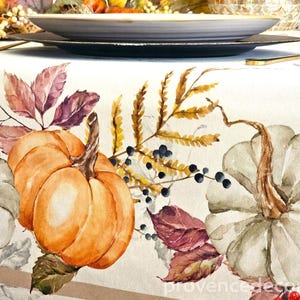 May include: A white tablecloth featuring a watercolor print of orange and pale green pumpkins, brown autumn leaves, and clusters of dark blue and red berries. The fabric has a smooth texture with a repeating seasonal botanical pattern.