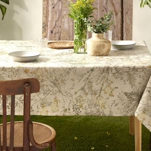 以下が含まれることがあります： ベージュ、緑、黄色の植物柄の長方形のテーブルクロスが木製のテーブルを覆っています。木製の椅子が前景にあります。テーブルには白いお皿、木製のボウル、花瓶が飾られています。