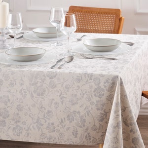 May include: A dining table set for a meal, featuring a floral patterned tablecloth in shades of gray and white. The table is set with white plates, bowls, silverware, and clear wine glasses. A lit candle in a glass holder adds to the ambiance.
