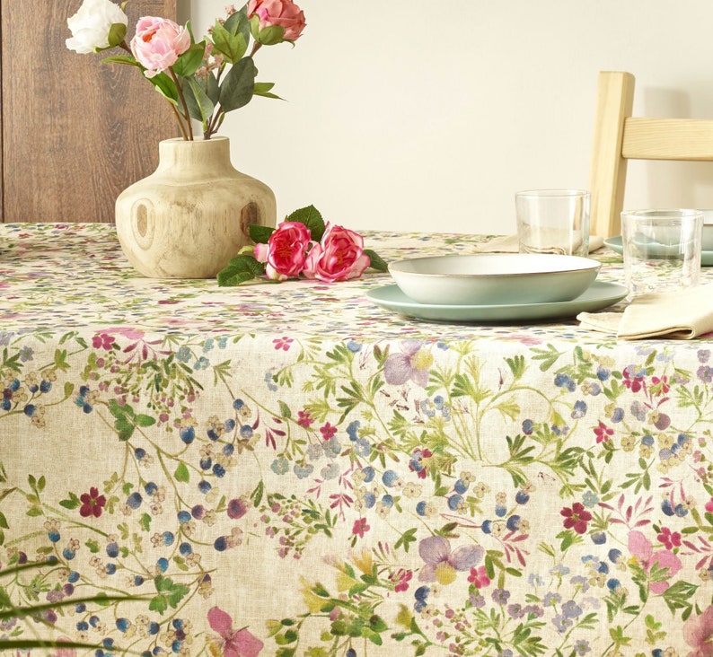 May include: A beige tablecloth with a floral pattern featuring pink, purple, and blue flowers. The tablecloth is set with a bowl, plates, and glasses.