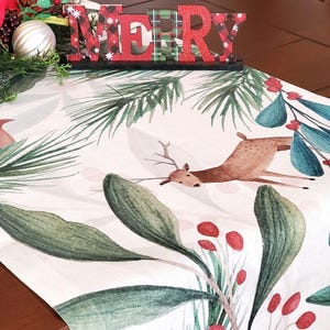 May include: A white Christmas table runner with a watercolor design featuring a deer, green leaves, and red berries. The runner is decorated with a red and green "MERRY" sign and festive greenery.