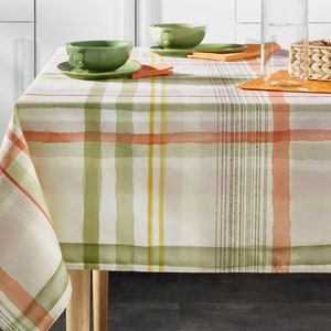May include: A white tablecloth with a green, orange, and yellow plaid pattern. The tablecloth is laid on a table with two green teacups and a glass pitcher.