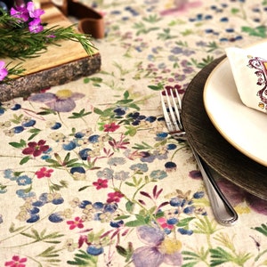 May include: A floral tablecloth with a cream background and a pattern of blue, purple, and red flowers. A white plate with a decorative napkin and a wooden charger sit on the tablecloth. A silver fork is placed next to the plates.