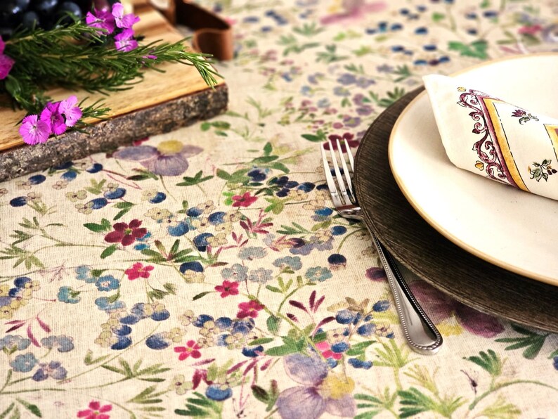 May include: A floral tablecloth with a cream background and a pattern of blue, purple, and red flowers. A white plate with a decorative napkin and a wooden charger sit on the tablecloth. A silver fork is placed next to the plates.