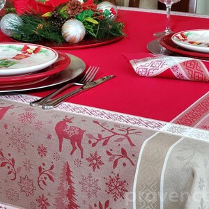 May include: Red and beige tablecloth with a white border and a snowflake and reindeer pattern. The tablecloth is set with a red plate, a white plate, and a red napkin.