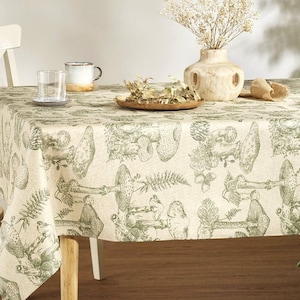May include: A beige linen tablecloth with a green mushroom and fern pattern. The tablecloth is laid on a wooden table with a vase of white flowers and two glasses.