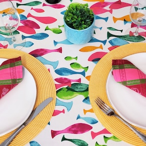 Peut inclure: Deux couverts avec des assiettes blanches, des sets de table en osier jaune et des serviettes rouges et roses avec des bordures vertes. Les couverts sont sur une nappe blanche à motif de poissons colorés.
