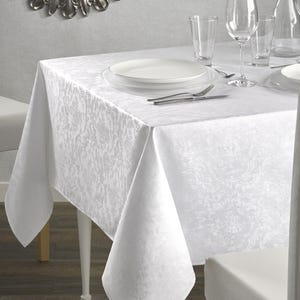 May include: A white tablecloth with a subtle floral pattern covers a table set for a meal. The table is set with white plates, silverware, and wine glasses. The tablecloth drapes over the edges of the table.