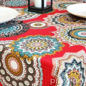 Puede incluir: Un mantel rojo con un detallado patrón de mandala circular en turquesa, dorado y negro. Platos blancos y cubiertos están colocados sobre la mesa, con una linterna negra en el centro. El mantel está hecho de una tela texturizada.