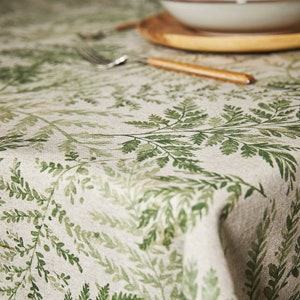 May include: A close-up of a beige linen tablecloth with a green fern pattern. The tablecloth is folded over on itself, showing the texture of the fabric.