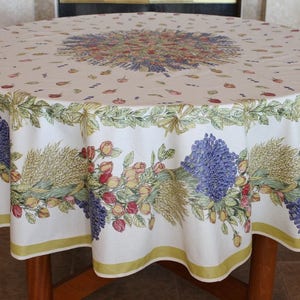 May include: A white round tablecloth with a floral pattern featuring lavender, wheat, and roses. The tablecloth has a green border with a scalloped edge.