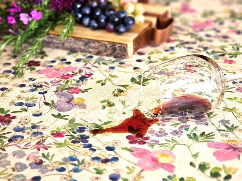 May include: A glass of red wine has been spilled on a white tablecloth with a floral pattern. The wine has created a dark red stain on the tablecloth.