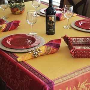 May include: A yellow and red tablecloth with a floral pattern. The tablecloth is set with red plates, wine glasses, and napkins with gold napkin rings. A bottle of wine is on the table, and a small red and white basket is in the center.