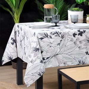 May include: White tablecloth with a black and white tropical leaf pattern. The tablecloth is laid on a table with black legs and a wooden surface. There are two white plates and two glasses on the table.