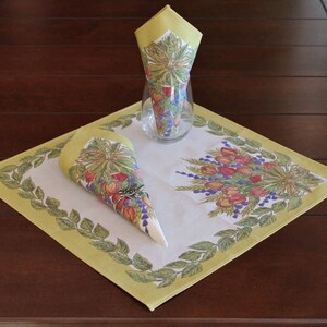 May include: A white linen table runner with a yellow border and a floral design. The runner features a green leafy border with a floral design in the center. Two yellow napkins with floral designs are folded into cones and placed on the runner.