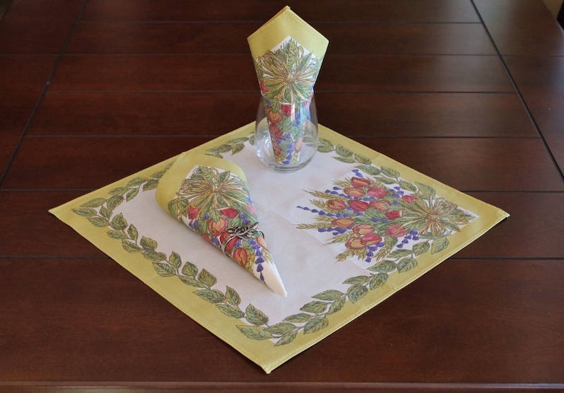 May include: A white linen table runner with a yellow border and a floral design. The runner features a green leafy border with a floral design in the center. Two yellow napkins with floral designs are folded into cones and placed on the runner.