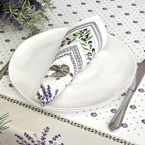 May include: A white ceramic dinner plate holding a folded white napkin with a lavender and olive branch print, secured by a silver-toned metal napkin ring. A silver fork and knife are placed on either side of the plate on a patterned tablecloth.