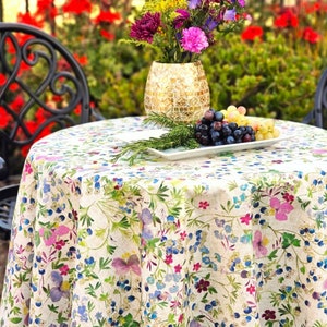 May include: A round tablecloth with a floral pattern featuring pink, blue, and purple flowers on a cream background. A white rectangular plate holds grapes and rosemary. A gold mosaic vase holds colorful flowers.